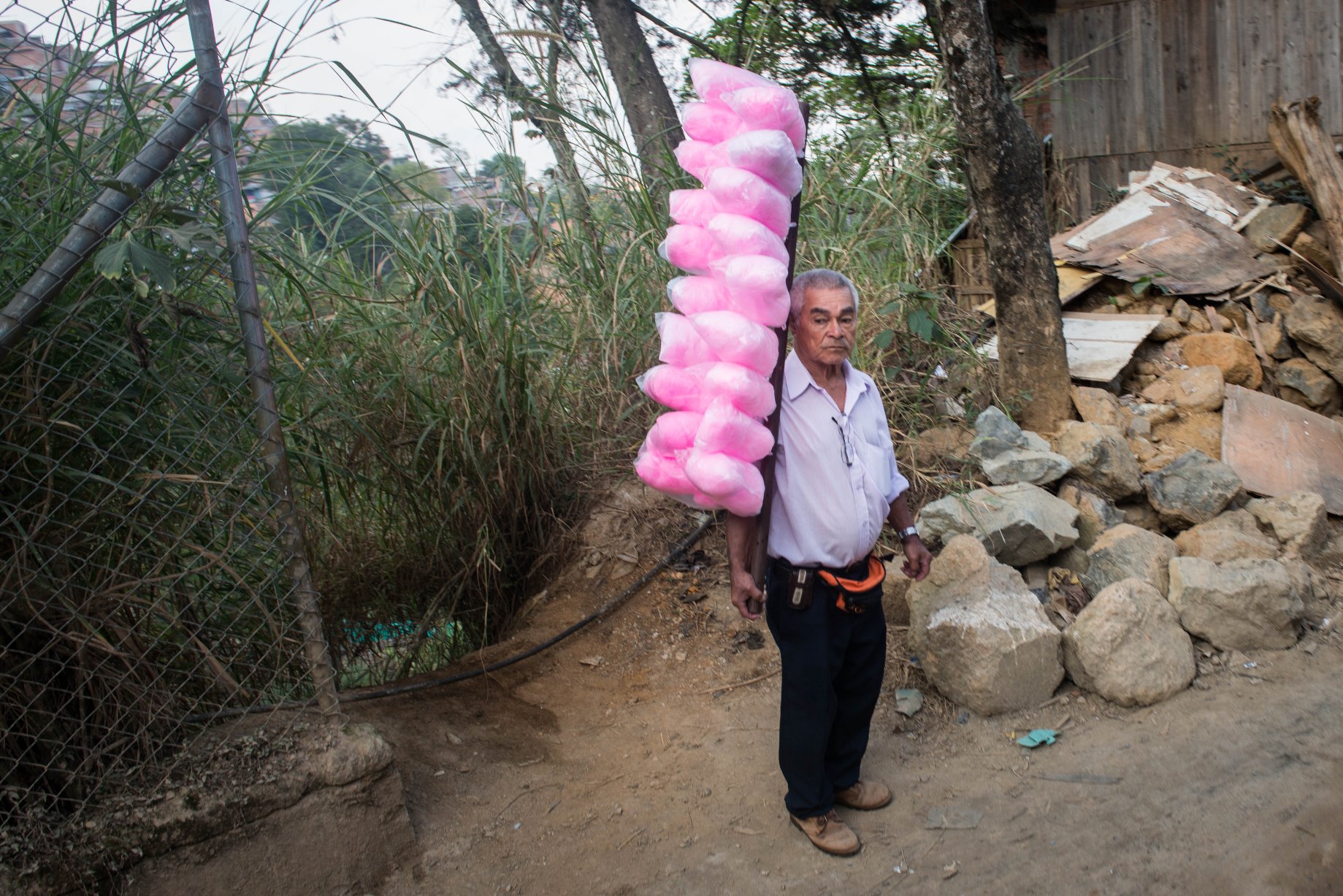 In the photograph, a man sells cotton candy on the roadside. Unemployment is a severe impediment to the community's development and well being. The local economy of Granizal remains in its infancy. Job markets in Bello or Medellin remain mostly inaccessible due to high transportation cost and limited access to educational and vocational training. The highest education attained by nearly half the population of Altos de Oriente is primary school with only 29% of the population attaining secondary education. Only a quarter of the community's working population engages in income generating activities, the majority of which is informal. Thanks to University of Antioquia's support, many community members are now receiving access to educational courses and programs. The younger generation of community members, however, are working hard to break these economic barriers. The availability of schools and relative peace have allowed those born and raised in Altos de Oriente to access schooling regularly. A number of the youth are the first in their families to attend college.